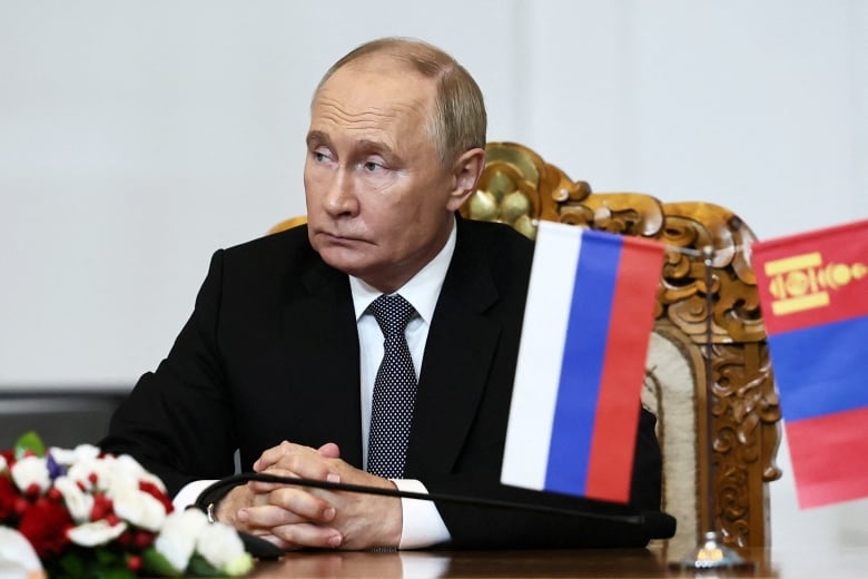 A stern man in a black suit with a black-and-white tie sits at a wooden table with his hands folded.