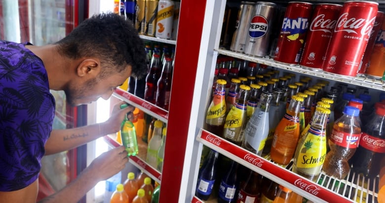 A man leans into an open beverage refrigerator, organizing cans and bottles of soda. 