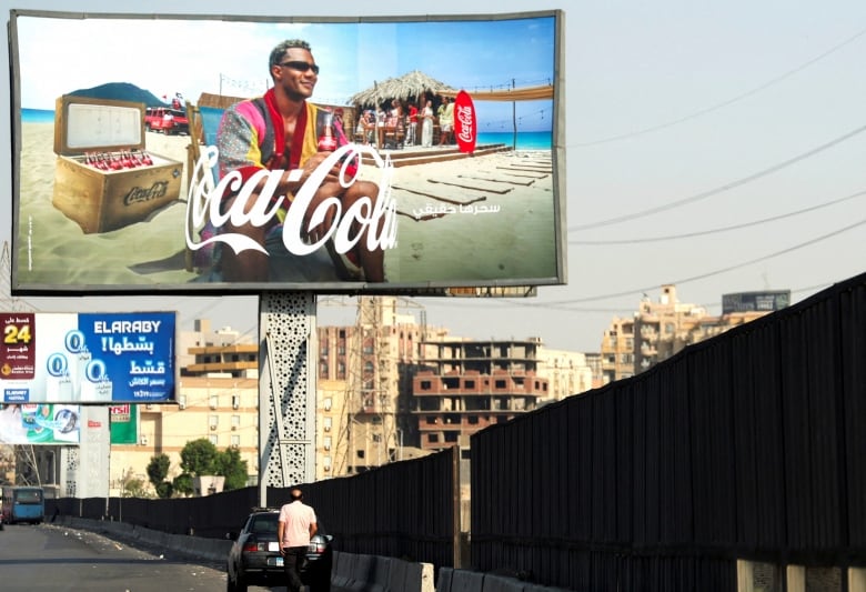 A billboard above a highway shows a man smiling underneath the Coca-Cola logo. 