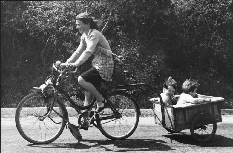 A black and white archive photo of a young mom biking with her children in  a  trailer