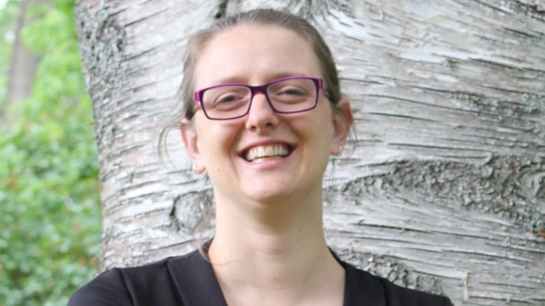 A portrait of a smiling woman wearing a black top and glasses, with her hair pulled back, standing in front of a birch tree.