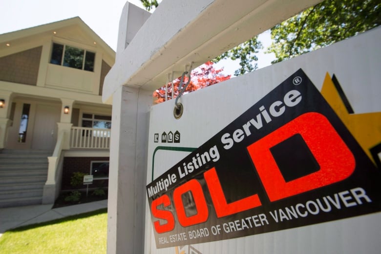 A 'for sale' sign stands on a lawn in front of a detached house. A sticker across it says 'sold.'