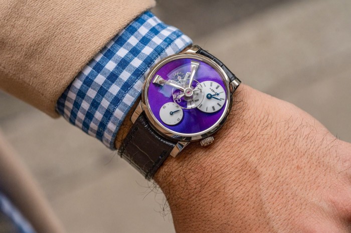 A luxury watch worn on a man’s wrist. The man is wearing a brown suit over a gingham shirt. The watch has a velvet coloured face and leather straps