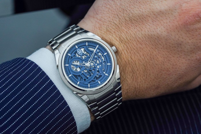 A luxury watch worn on a man’s wrist. The man is wearing a dark blue suit over a white shirt. The watch has a blue-coloured face and metal straps