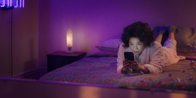 A still from a movie shows a woman holding a phone lying on a bed, in a room lit by purple light.