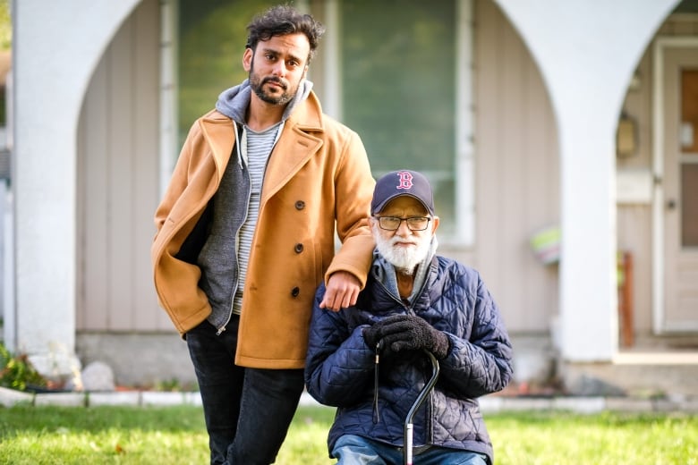 A younger man stands next to an older man seated outside a house.