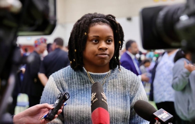 A young woman in a blue sweater stands in front of three sets of mics. 