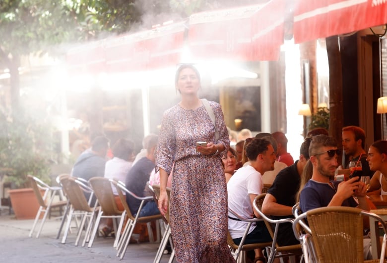 A woman in a floral dress walks by a bar where people are seated outside that's spraying a fine mist of water those seated outside and this woman walking by.