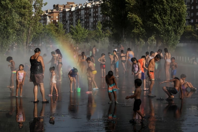 A crowd of people on city pavement are getting misted by water and treated to a rainbow. 