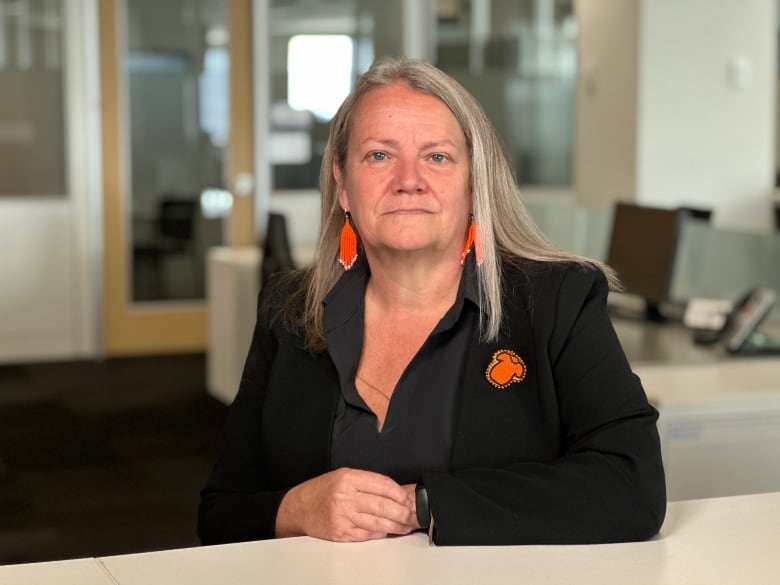 A person wearing black clothes and bright orange beaded earrings and a pendant sits at a desk in an office setting.