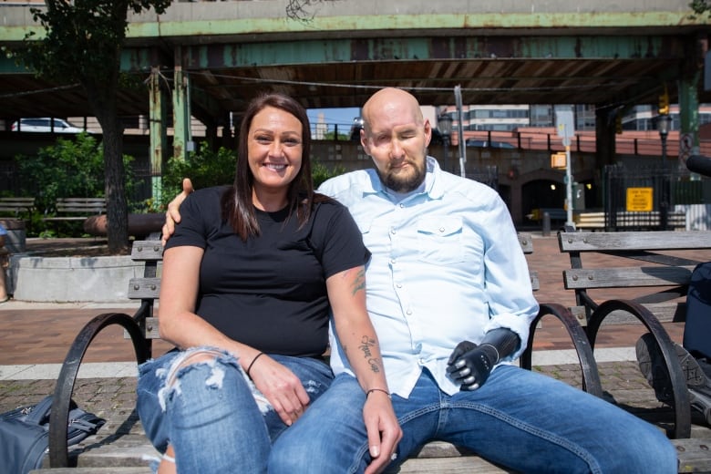 A man and woman sit together on a bench. The woman has her hand resting on the man's inner thigh, and he has his arm around her shoulder. His left eye is closed, and his left arm is a black, metallic prosthetic. 