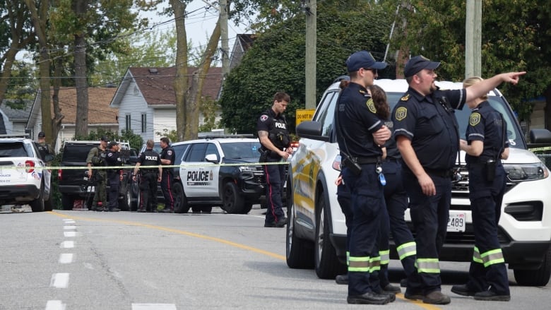 A group of paramedics stand in the foreground. Down the street is a circle of police officers surrounded by caution tape and cruisers.