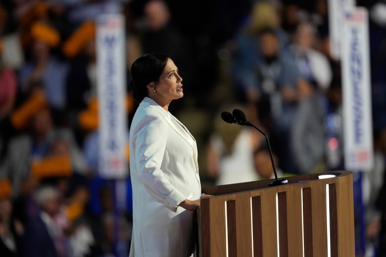 A brunette woman wearing a white blazer speaks at a podium.