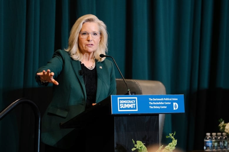 A woman with blond hair and glasses speaks at a podium. 