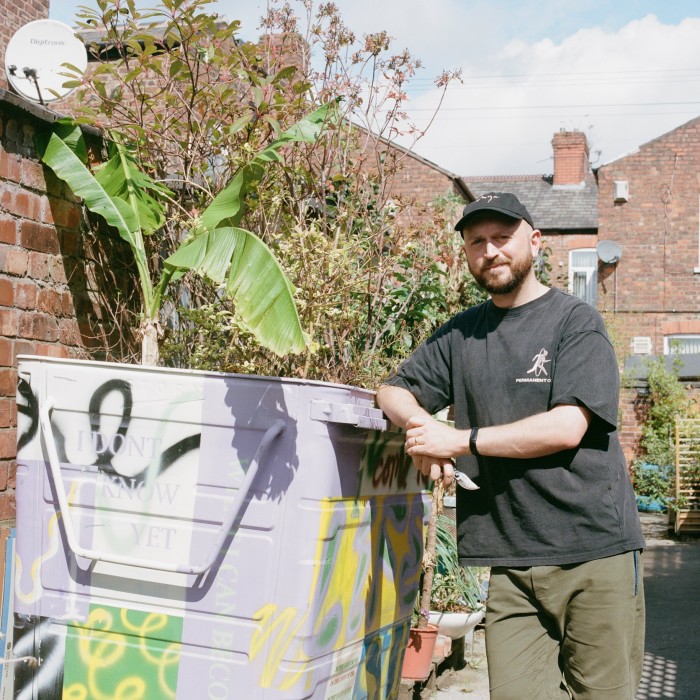 A man leans on a large metal wheelie bin, which has been painted lilac and is now being used as a giant flowerpot for plants