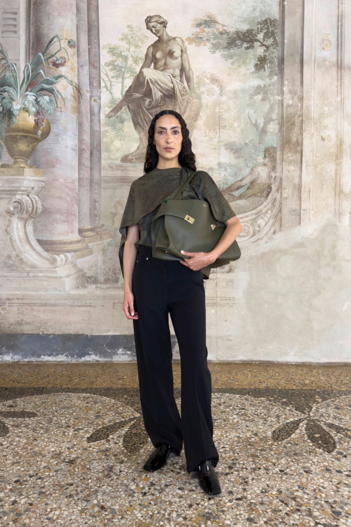 A dark-haired woman stands in front of a frescoed wall, wearing black trousers and grey blouse, and clutching a leather handbag under her arm