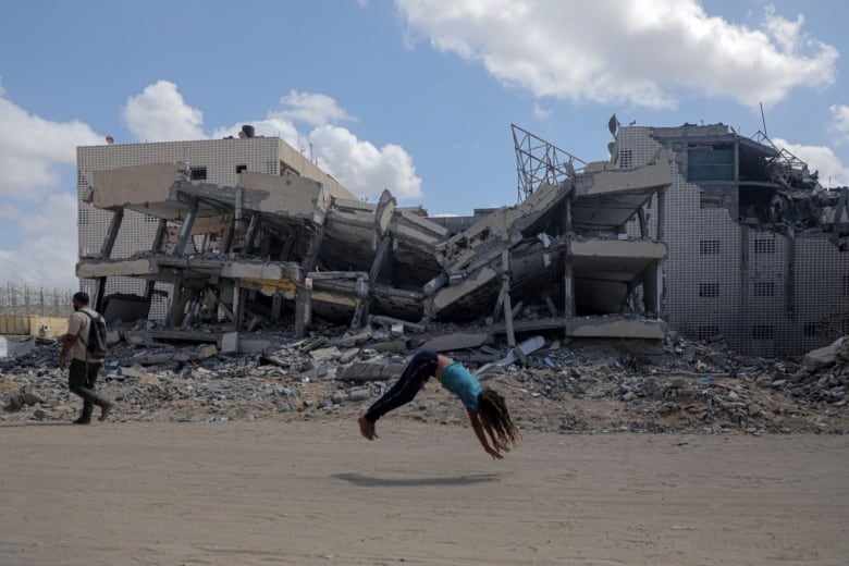 A child does a backflip in front of a destroyed building in a war-torn region.