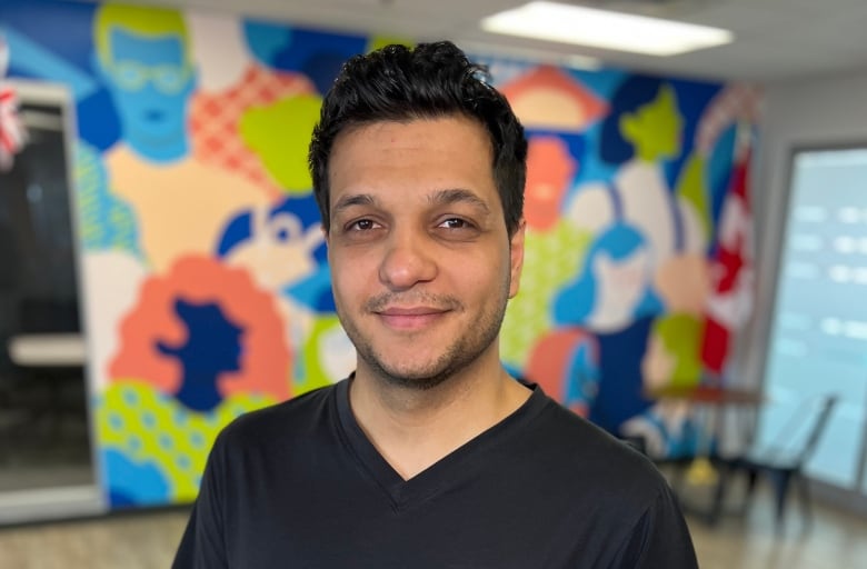 A smiling young man in a black t-shirt stands in an indoor room, with a colourful mural seen behind him.