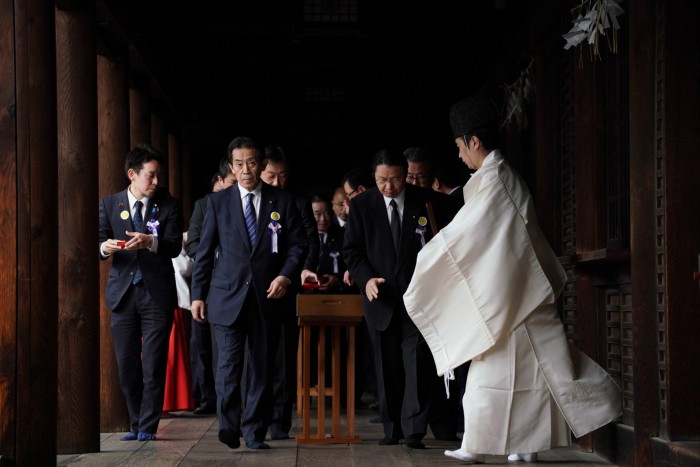 Japanese lawmakers, wearing suits and purple ribbons, visit the Yasukuni Shrine at the end of the spring festival in Tokyo on April 23 2024. A Shinto priest in traditional white robes stands to the right, guiding the group.