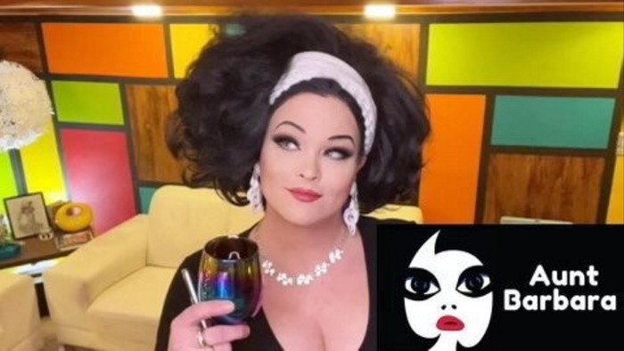 Jennifer Suchan, also known as Aunt Barbara, poses with a drink in hand. She is wearing a black dress, a white headband, and large earrings. The background features a colorful, retro-style wall and a cream-colored couch. The bottom right corner of the image includes a logo with a stylized face and the text ‘Aunt Barbara’