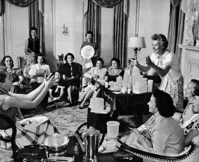 Brownie Wise enthusiastically demonstrates a Tupperware bowl filled with water to a group of women at a Tupperware Party in the 1950s. The women are gathered in a living room, seated around a table displaying various Tupperware products.