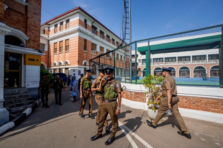 Half a dozen police officers in green uniforms stand together on the street on a sunny day.