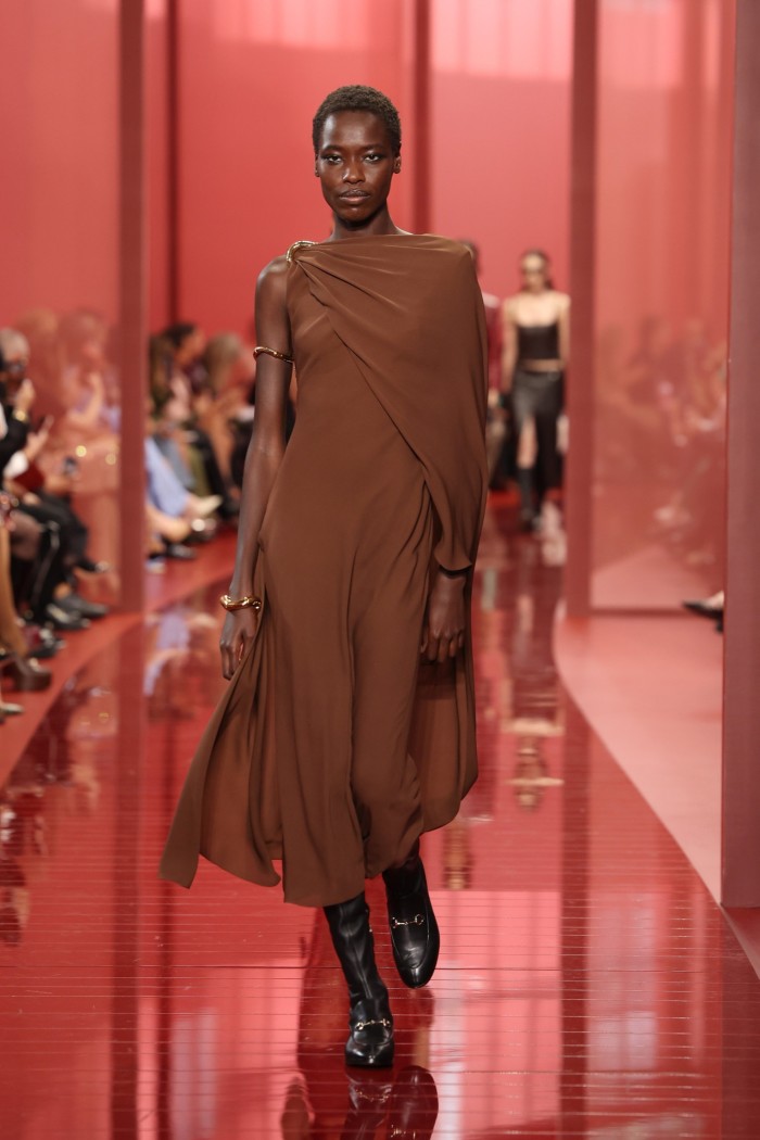 A model on a red catwalk wears a draped, flowing brown gown