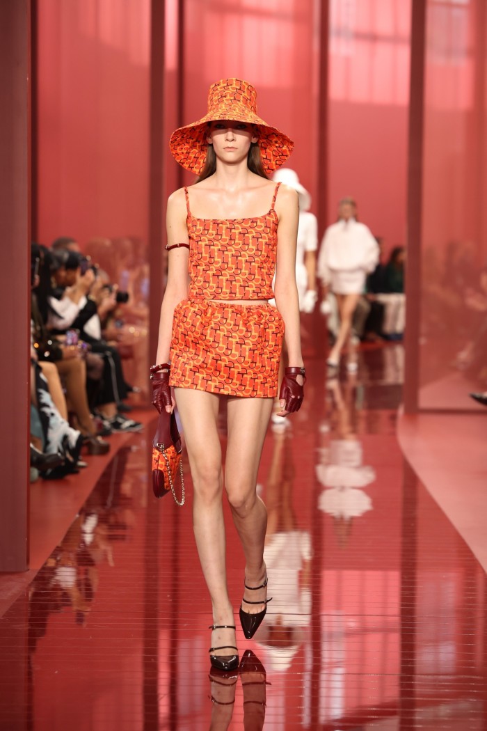 A female model on a red catwalk wearing a bright orange mini-skirt and wide-brimmed hat of similar colour