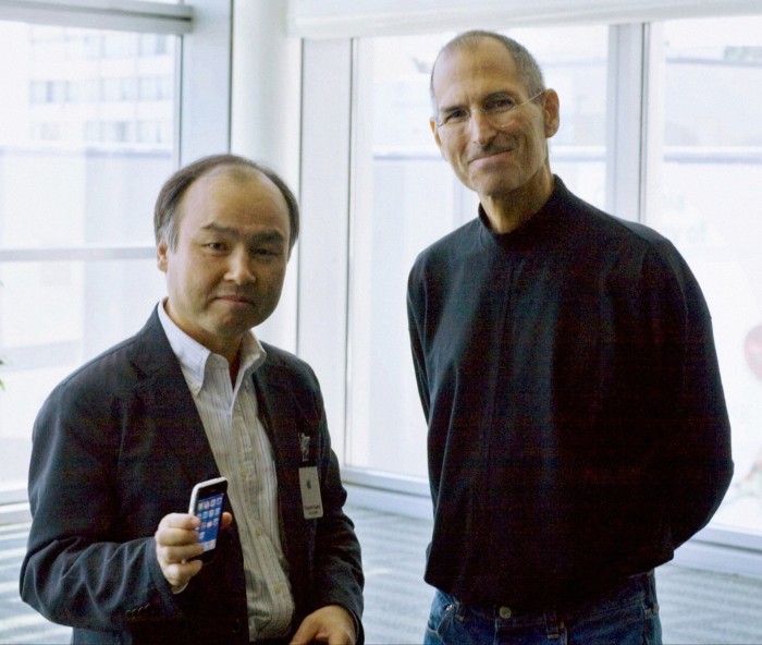 Two men stand smiling. Son, the shorter man, wearing open-neck shirt and jacket, holds a mobile phone. Next to him, Jobs, taller, wearing black rollneck top and jeans, stands with his hands behind his back