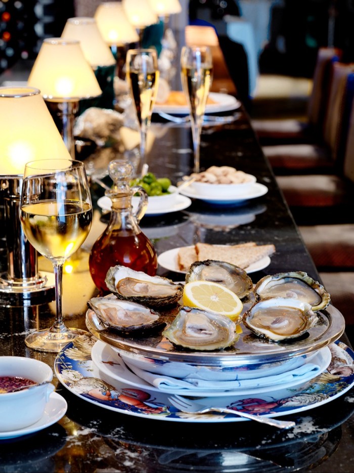 A platter of oysters sits at the center of a lavishly set bar table, accompanied by a glass of white wine, a small carafe of sauce, and other appetizers. The background features champagne flutes and glowing lamps