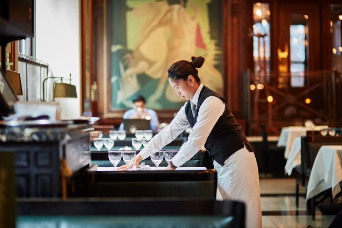 A focused waiter prepares a table in an upscale restaurant, placing glassware neatly on a white tablecloth. The background features a large mural and wooden decor