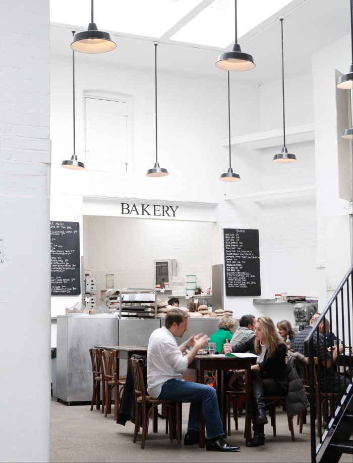 A bright bakery café with high ceilings and hanging pendant lights. Customers are seated at wooden tables, engaging in conversation, while the bakery counter in the background displays fresh bread. Chalkboards list menu items on the white brick walls