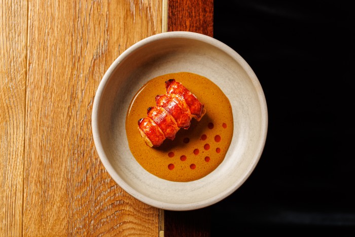 A neatly presented lobster tail served in a shallow bowl with a rich orange-brown sauce, accented with small red oil droplets. The dish is placed on a wooden table