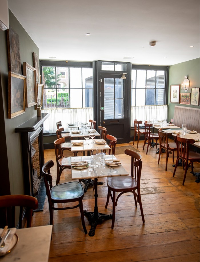 A dining area featuring marble tables and wooden chairs on a warm wooden floor. Large windows with short curtains bring in daylight, while the walls are decorated with framed paintings