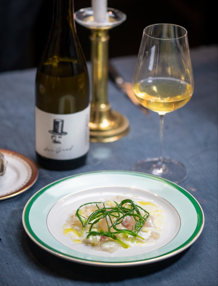 A fine dining plate of seafood garnished with green herbs and olive oil, accompanied by a glass of white wine