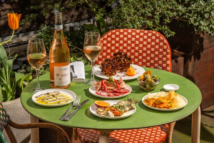 A green round table in a garden setting, offering a spread of Mediterranean appetizers including sardines, charcuterie, grilled artichokes, and roasted vegetables, alongside a bottle of rosé and two wine glasses