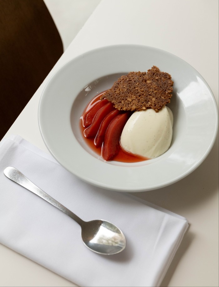 A dessert plate featuring poached pear slices in a red sauce, accompanied by a scoop of vanilla ice cream and a crispy almond tuile on top. The dish is served on a white plate with a spoon and napkin beside it