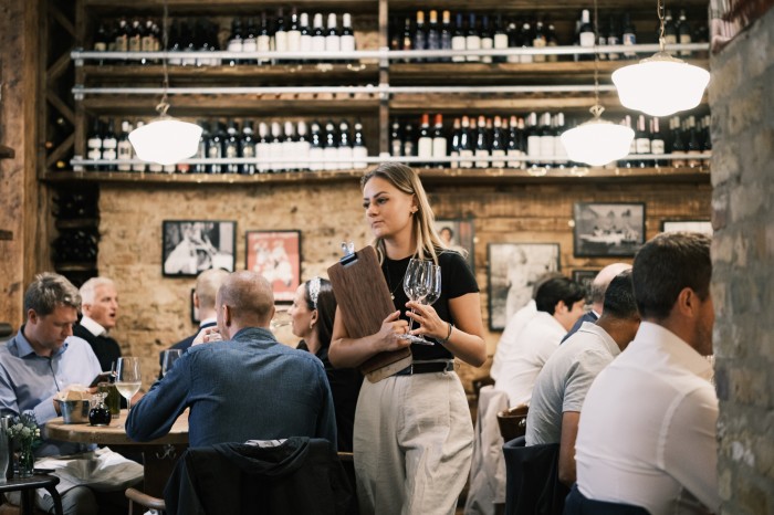 A busy restaurant scene with a waitress holding wine glasses and a clipboard, navigating through a crowd of seated patrons. The rustic interior features shelves of wine bottles, wooden walls, and framed artwork