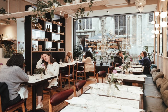 A bright, modern café with several groups of people sitting at tables, talking and dining. The décor includes hanging greenery, minimalistic shelves, and large windows letting in natural light from the street view outside