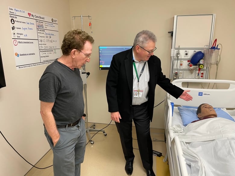 Two men stand in a hospital room where a simulated patient dummy is on a hospital bed.