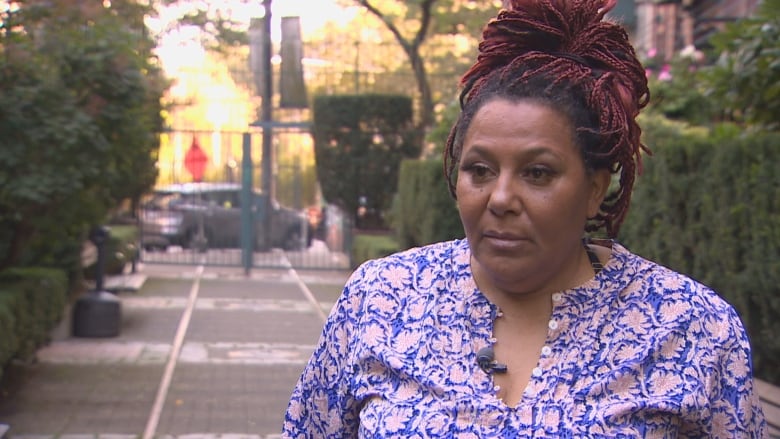 A Black woman wearing a purple and pink top speaks next to a hedge.