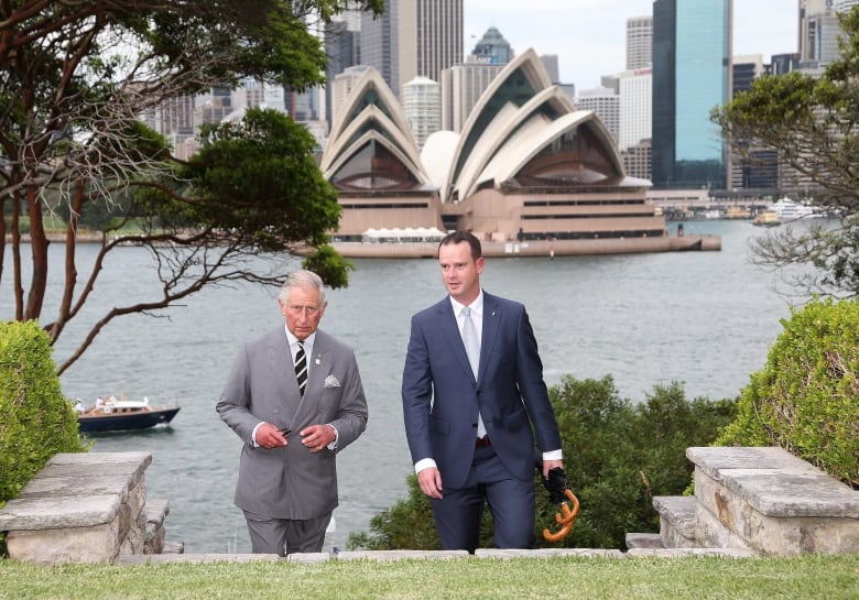 Two people walk up stone stairs toward a grassed area with a body of water and the Sydney Opera House in the background.