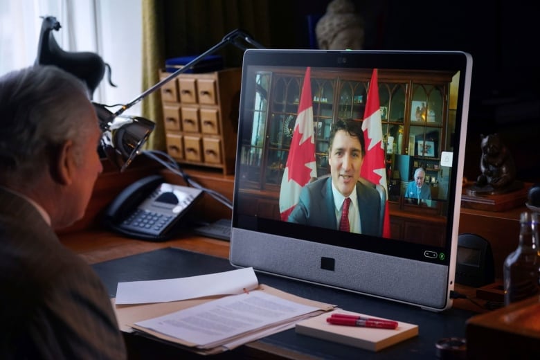 A person sitting at a desk speaks with another person via a video call, with that person's face visible on a monitor.