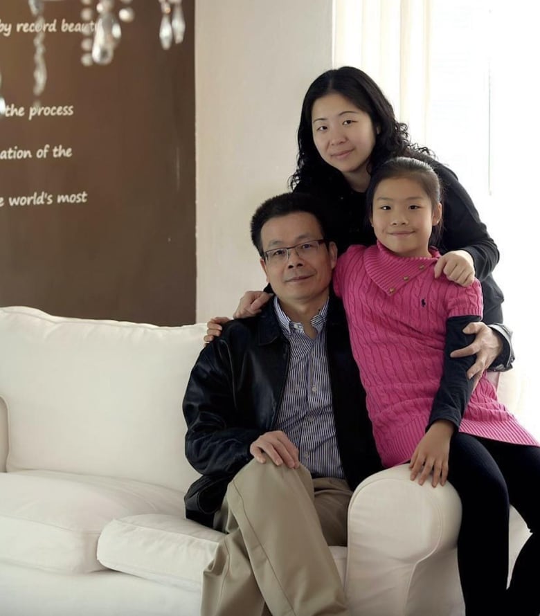 A smiling girl sits on a couch armrest. Next to her is a smiling man sitting on a white couch. A smiling woman stands behind them both with her arms around their shoulders.  