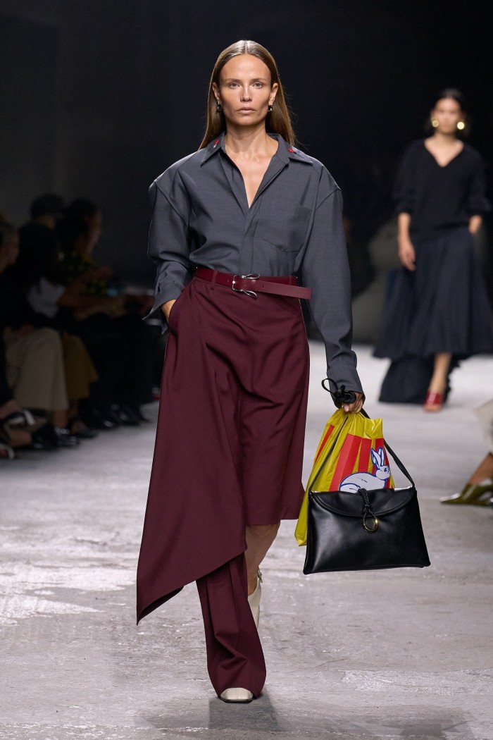 A female catwalk model wearing a silky dark open-neck shirt with a maroon-coloured half-skirt and half-trouser and carrying a handbag decorated with a rabbit motif