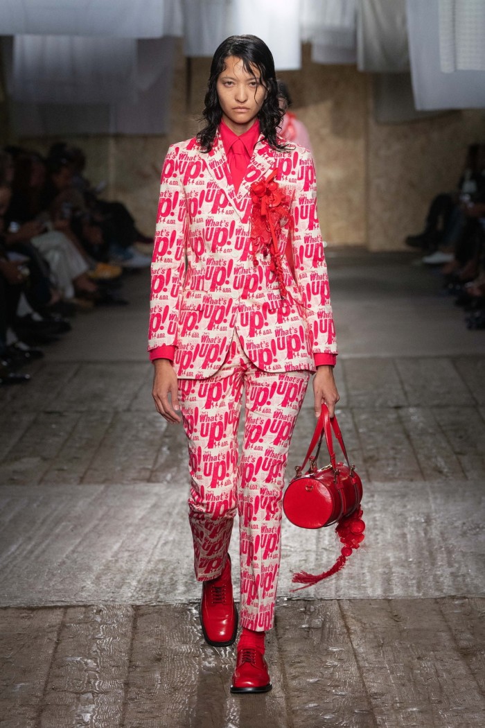 A female catwalk model wearing a trouser suit in white covered all over with the words ‘what’s up’ in red. Her shoes, handbag, rosette and shirt and tie are also bright red