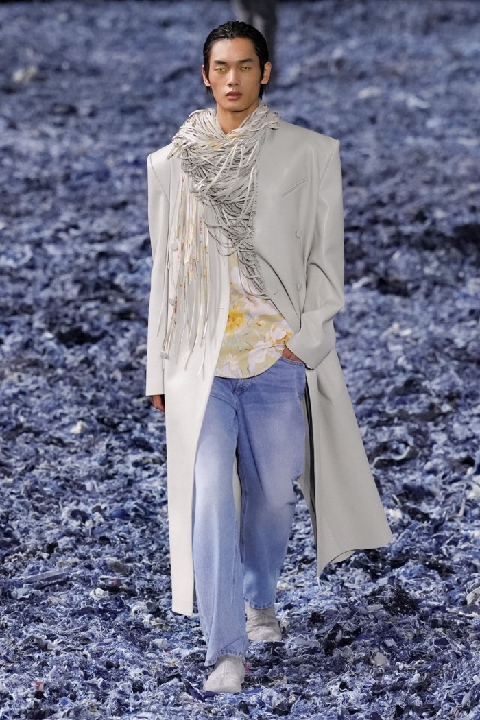 An androgynous catwalk model with striking blue contact lenses wearing a long pale grey coat, baggy faded jeans and striding across a floor carpeted with strips of denim 