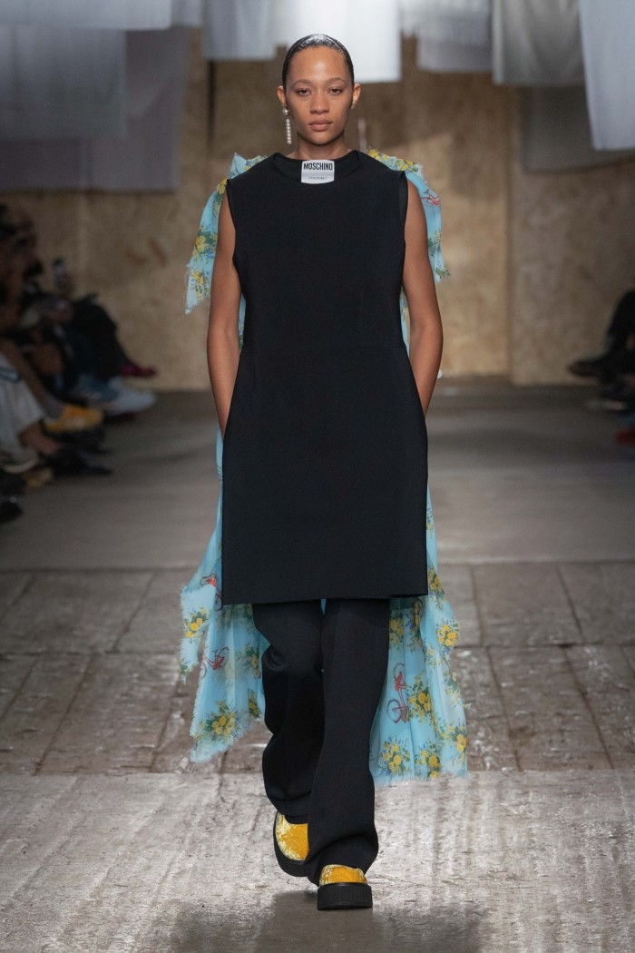 A female catwalk model in a minimalist combo of sleeveless skirt and trousers in plain black — but with the word ‘Moschino’ at her neck and a blue floral-patterned dress only partly visible stitched to her back