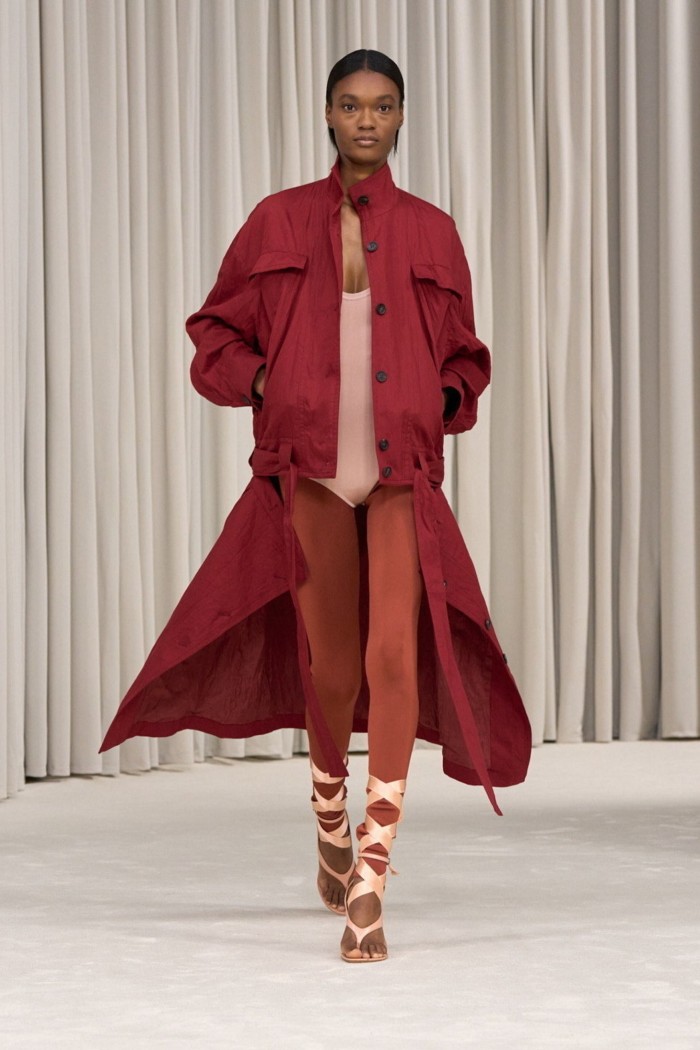 A female catwalk model wearing a long red trench-style coat, open to reveal a ballet dancer-style leotard, stockings and laced-up pumps