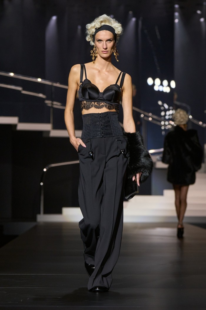 A female catwalk model with Madonna-like blond hair or a wig, wearing a silky black lacey bra and wide high-waisted black trousers